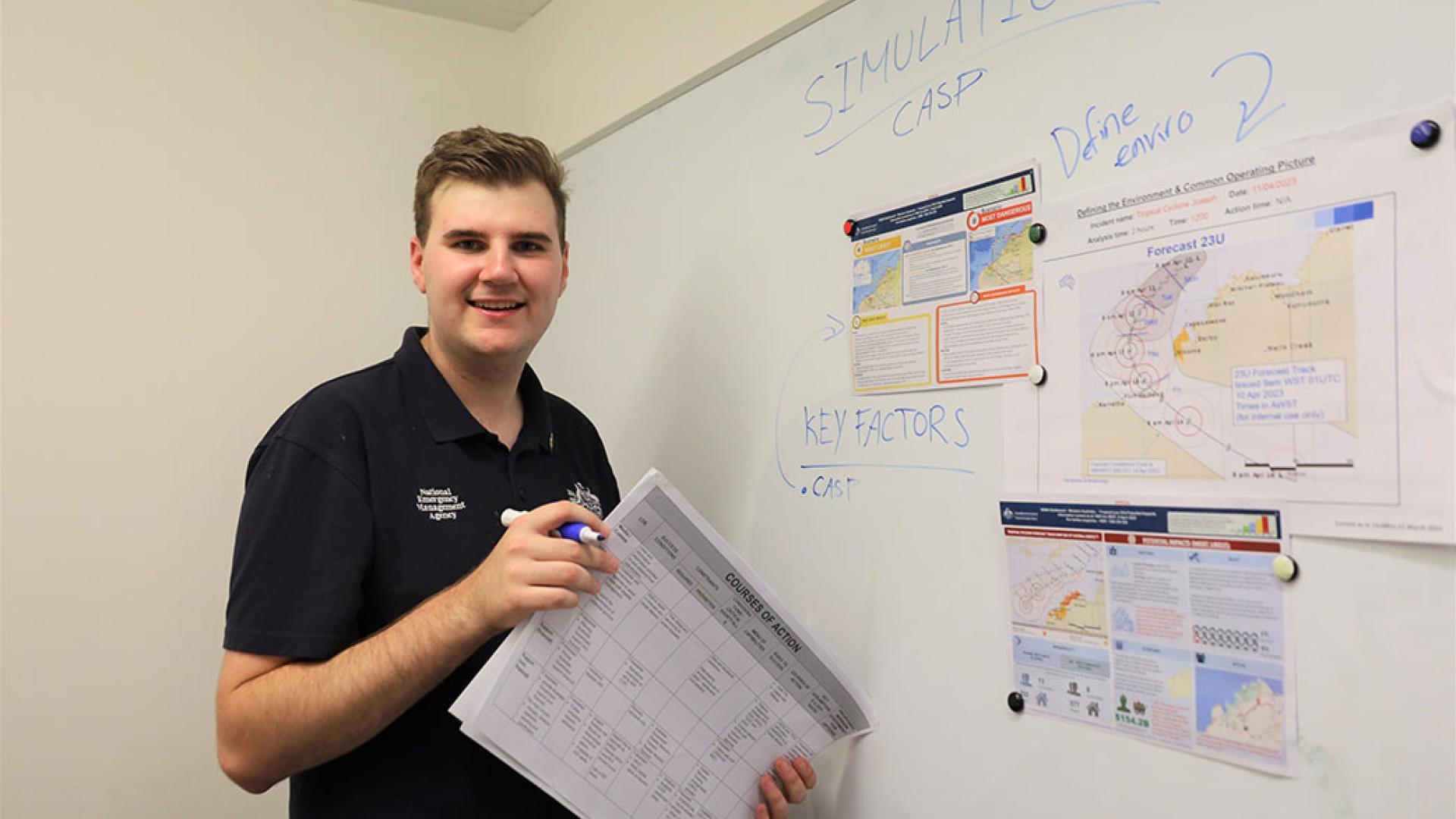 A person in a navy polo shirt with NEMA branding standing on the left of a whiteboard. They’re holding a whiteboard marker and a paper folder and are smiling. 
