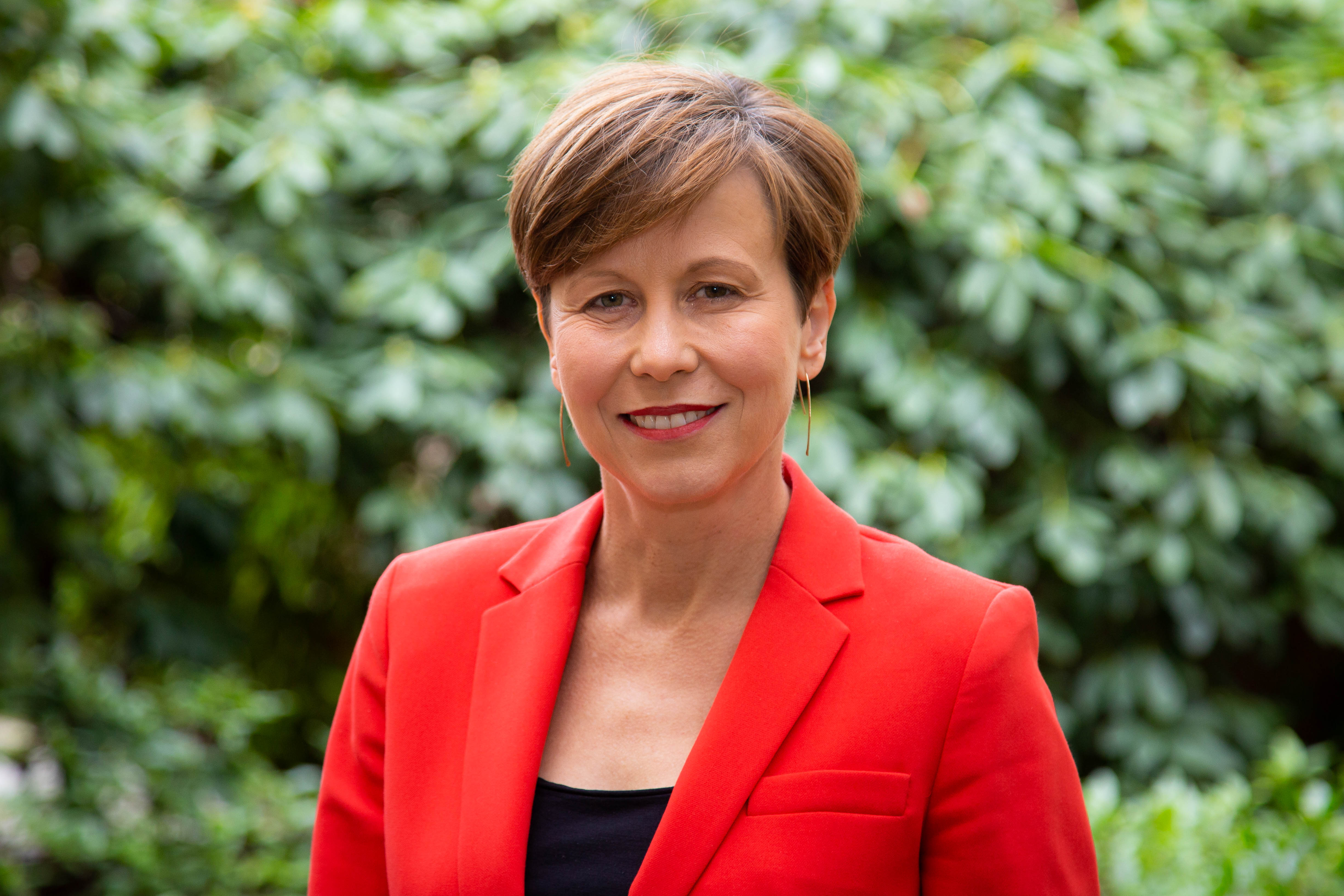 A woman wearing a red blazer poses for a portrait outside in front of green plants.