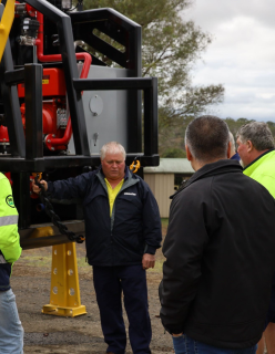 A man with grey hair stands beside a large tractor, while conversating with four other men who are dressed in high vis outfits.  