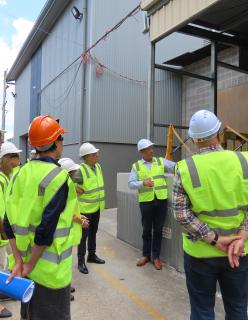 People wearing high-visibility protective vests and hard hats stand together outside a large warehouse.