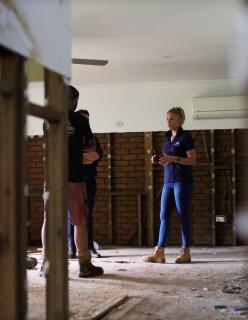 A NEMA employee in a blue outfit and work boots talks to two people in a disaster impacted room with exposed wooden beams and brick walls.
