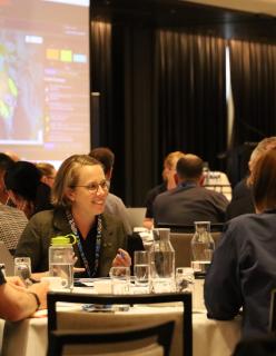 People sit in a conference room speaking at a table. A map of Australia is displayed on a large screen behind them.