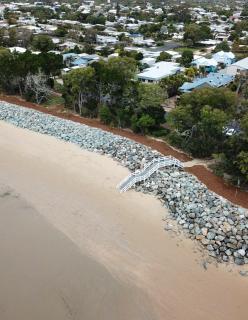 Aerial photo of a beach with new rock wall, footbridge over it, and behind a line of trees are roofs of houses.