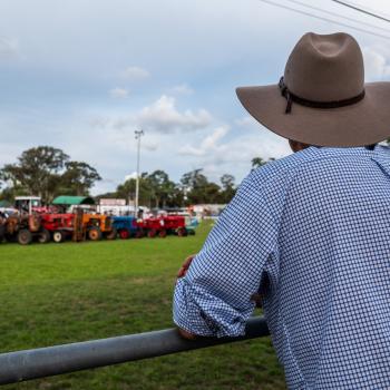 A farmer leans on a fence post overlooking tractors.