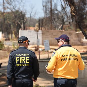 Two fire personnel in uniform walking outdoors, surrounded by trees and open ground under a clear sky. On the back of their uniforms reads ”Country Fire Service” and the other ”NSW Rural Fire Service”.