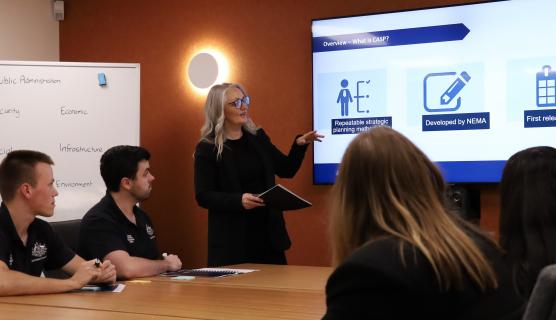 A presenter points to a large screen while people sit at a meeting table listening to the presentation.