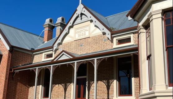 A historic building featuring a porch under a clear blue sky, surrounded by grass and plants, showcasing architectural details like sash windows and siding.