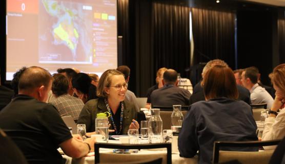 People sit in a conference room speaking at a table. A map of Australia is displayed on a large screen behind them.