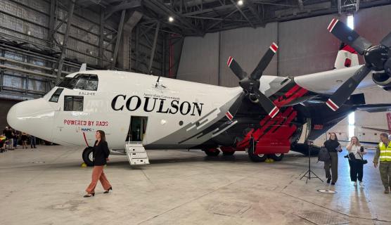 Inside a large aircraft hangar, side view of a large plane with 2 propellers on the wing, 'Coulson' and the Australian Government logo on the side. Minister McBain is walking past and some media and a person in a hi-vis vest are to the side.