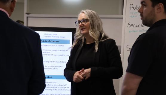 Photo of Bec, hands folded, talking to 2 other people in an office with a whiteboard and large screen behind her.