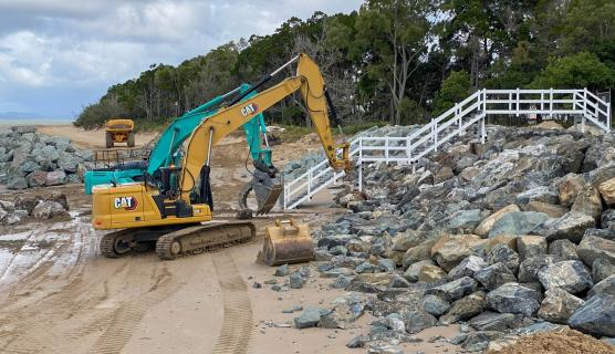 A sea wall under construction. Two large diggers are moving large grey rocks on the edge of the sand, with green bush in the background.