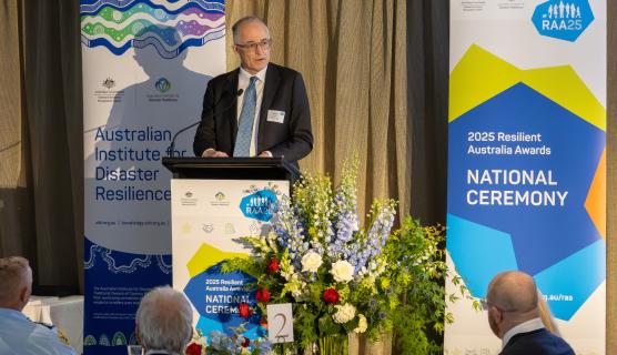 Man in a suit and tie speaking at a lectern with banners behind him that say 'Australian Institute for Disaster Resilience' and '2025 Resilient Australia Awards National Ceremony'. In the foreground are a few people seated at table 2 with a large floral arrangement in the middle of the table.