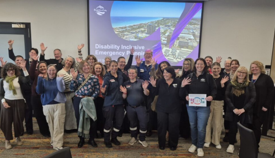 Large group of 25 people posing for a group photo, most with their hands in the air. In the background is a large screen with 'Disability Inclusive Emergency Planning' on a slide.