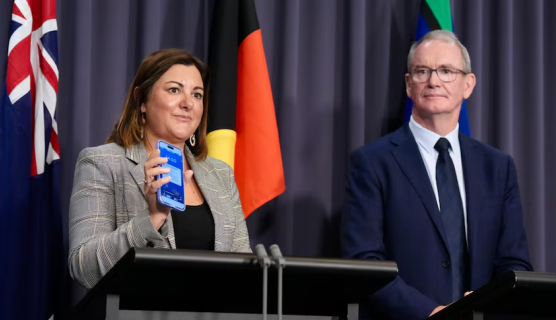 Photo of Minister Kristy McBain standing at a lectern holding up a mobile phone that has an alert on the screen. Next to her is Brendan Moon in a suit with tie standing at a second lectern. Behind them are the Australian, Aboriginal and Torres Strait Islands flags.