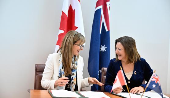 Photo of two women sitting at a desk smiling and talking to each other, signing papers in folders, with Canadian and Australian flags behind them.