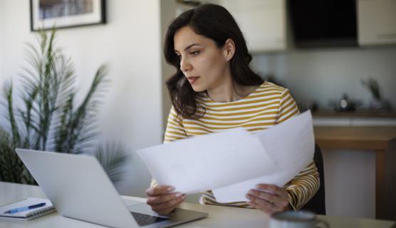 Woman sitting at a table looking slightly concerned at a laptop with papers in hand.