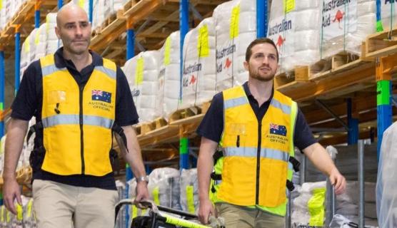Two men carrying a generator between them, wearing high vis yellow vests, walking in a warehouse with tall stacked shelves behind them of packages labelled 'Australian Aid'.