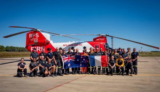 Large group of people posing in front of a large red and white RFS helicopter, holding large Australian and French flags, on a concrete runway area with blue sky.