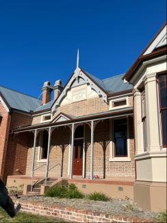 A historic building featuring a porch under a clear blue sky, surrounded by grass and plants, showcasing architectural details like sash windows and siding.