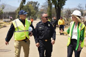 A group of three wearing safety vests and hard hats walking together on a road, surrounded by trees. A second group comprised of three is in the background.