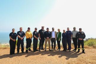 A group of emergency management experts standing together outdoors on a hilltop overlooking the ocean, wearing various clothing and footwear, with a clear sky in the background. 