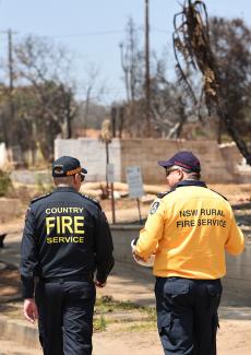 Two fire personnel in uniform walking outdoors, surrounded by trees and open ground under a clear sky. On the back of their uniforms reads ”Country Fire Service” and the other ”NSW Rural Fire Service”.