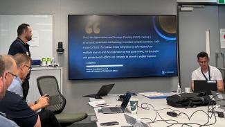 Group of men with laptops sitting around a table. One man is presenting to a slide with a quote from Joe Buffone (Deputy Coordinator General, NEMA) that says 'The Crisis Appreciation and Strategic Planning (CASP) employs a structured, systematic methodology to analyse complex scenarios. CASP is a set of tools that allows timely integration of information from multiple sources and the exploration of how government, not-for-profit and private sector efforts can integrate to provide a unified response.