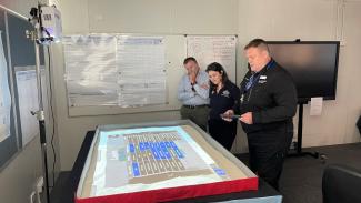 3 people standing over a table with a map and data table projected on it. One person is holding a tablet to control what data shows on the projection.