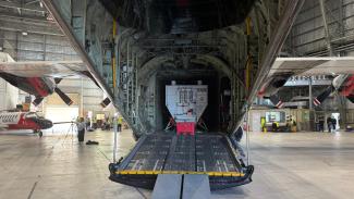 Rear view of a large plane with its rear hatch open to show the inside of the fuselage with lots of cables and frame visible, and a large square tank in the middle.