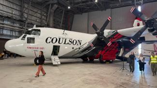 Inside a large aircraft hangar, side view of a large plane with 2 propellers on the wing, 'Coulson' and the Australian Government logo on the side. Minister McBain is walking past and some media and a person in a hi-vis vest are to the side.