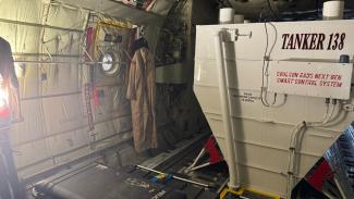 Inside a large plane with the large square tank to the right that reads 'Tanker 133'. To the left you can see the curvature of the fuselage, a closed door with a small round window, and a uniform/over-suit hanging up.