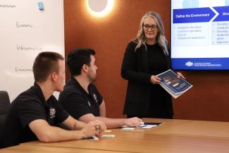Bec presenting in a board room in front of a ppt, holding a printed document.