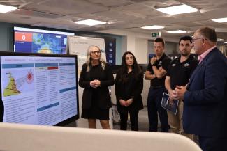 Group of 5 NEMA staff, including Bec and Joe Buffone, in the National Situation Room discussing a dashboard that's tracking a cyclone approaching Queensland.