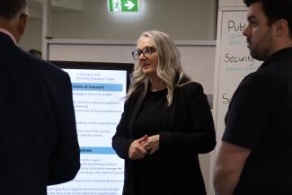 Photo of Bec, hands folded, talking to 2 other people in an office with a whiteboard and large screen behind her.