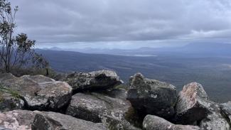 A high point view across the Grampians with large grey granite rocks in the foreground, a scraggly shrub to the left, and bushland in the distance with mountains in the far distance, a body of water and cloudy sky.