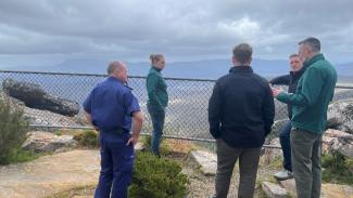 5 people standing casually. 1 person looks to be explaining something. There's a chest height wire fence at the edge of the lookout, with large grey granite rocks on either side and the mountain range in the distance with clouds overhead.