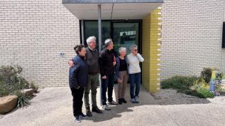 5 people standing in a row, posing for a photo off-camera, in front of a pale-coloured brick building with glass door.