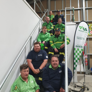 Group photo of a rescue team on the stairs inside the centre, wearing green and navy coloured uniforms with a 'VRA Rescue NSW' banner to the side.
