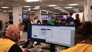 Photo of the National Situation Room: in the foreground, 2 people wearing yellow vests looking at large monitors (which are blurred out) and in the background a group of people gathering in what looks like a briefing, wearing different coloured vests (grey, green, navy).