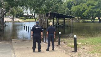Photo from behind 2 people (one with 'Police' on their back) looking at a flooded park with a barbecue pavilion half submerged in the middle.