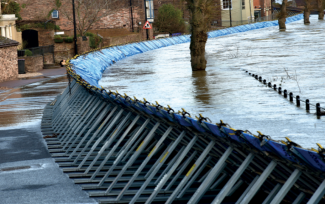 A flood barrier propped up from the left with many angled rods, with floodwater flowing on the right side and some tree trunks and the top of a fence visible in the water.