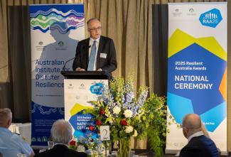 Man in a suit and tie speaking at a lectern with banners behind him that say 'Australian Institute for Disaster Resilience' and '2025 Resilient Australia Awards National Ceremony'. In the foreground are a few people seated at table 2 with a large floral arrangement in the middle of the table.