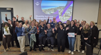 Large group of 25 people posing for a group photo, most with their hands in the air. In the background is a large screen with 'Disability Inclusive Emergency Planning' on a slide.
