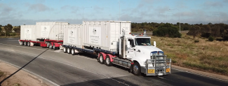 A large road train truck on a highway with bush in the background, with 4 white containers on its 2 trailers. The containers have the Australian Government crest logo on them. 