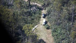 Aerial photo of the bush with a clearing and a fire trail road leading away from it. There are 2 vehicles and some large white boxes/packages along the left of the clearing.