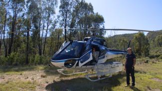 Small helicopter in a grassy clearing with blue skies and bush in the background, one person inside the helicopter with the door open and a man standing outside next to it.