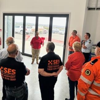 Group of SES volunteers in orange or black with orange logo, standing inside at large glass doors with a big red ribbon across them.