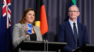 Photo of Minister Kristy McBain standing at a lectern holding up a mobile phone that has an alert on the screen. Next to her is Brendan Moon in a suit with tie standing at a second lectern. Behind them are the Australian, Aboriginal and Torres Strait Islands flags.