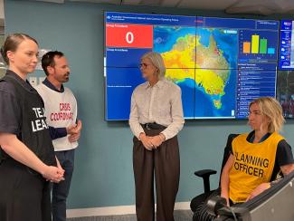 Phot of Dave Long wearing a white vest with red text that says Crisis Coordinator talking to the Governor General Sam Mostyn. Another team member woman stands nearby wearing a black Team Leader vest, and a third woman is sitting to the right and wearing a yellow Planning Officer vest. Behind them is a screen with a map of Australia showing no current emergencies.