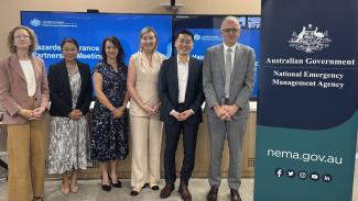 4 women and 2 men dressed professionally, standing in a line next to a pull-up banner that has the Australian Government crest and National Emergency Management Agency.
